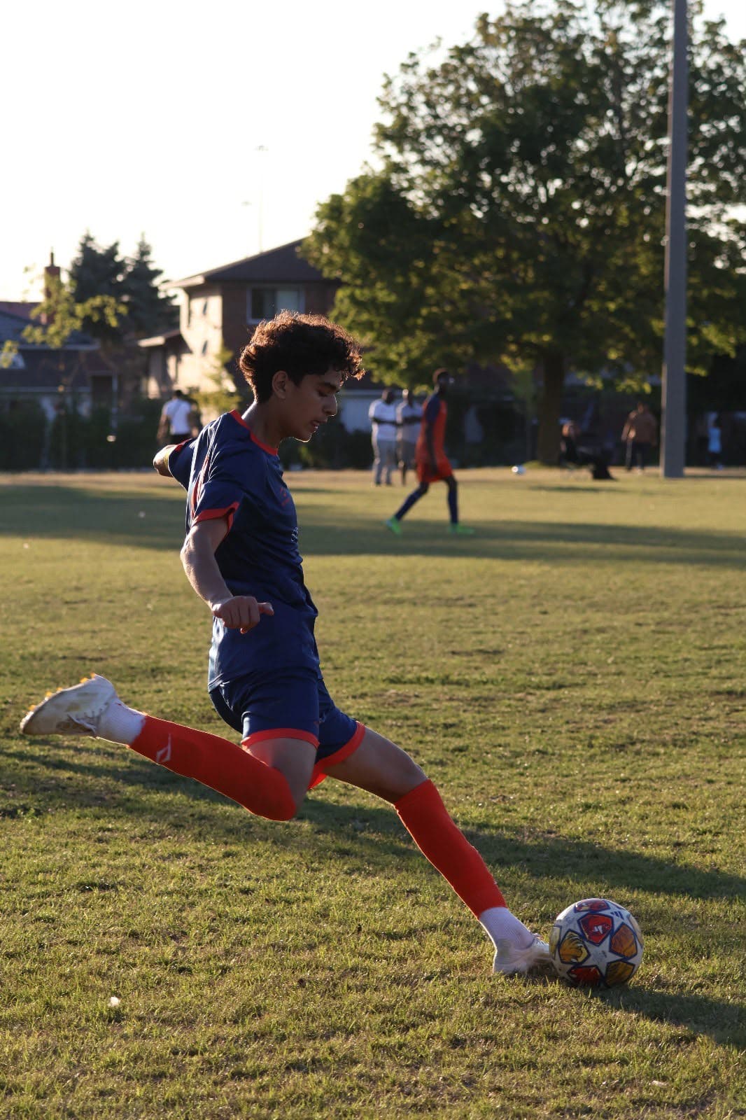 A young footballer drives a ball across a Toronto grass field in late-afternoon sunlight