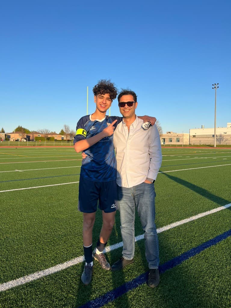 A young football captain stands with a family member on an artificial turf pitch in Toronto